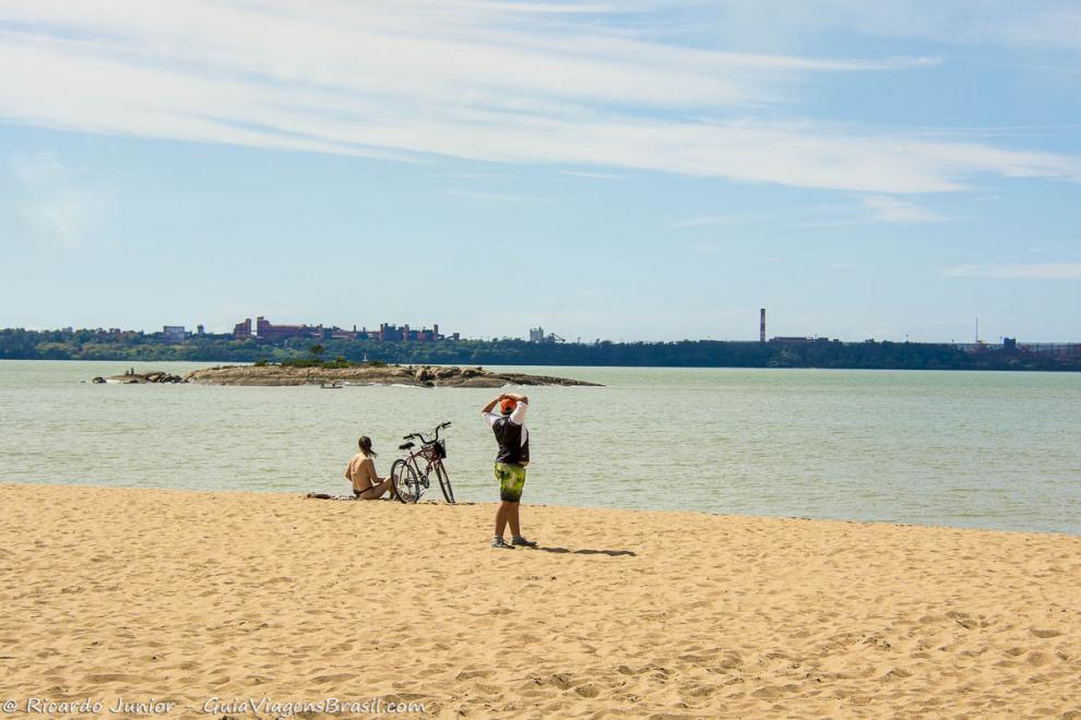 Imagem de uma menina sentada na areia com sua bicicleta ao lado e um rapaz se espreguiçando.